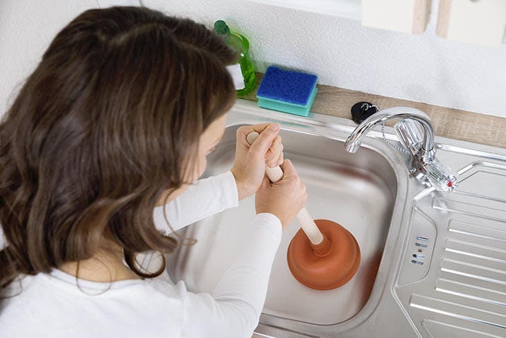 A woman unclogs a stainless steel sink with a plunger